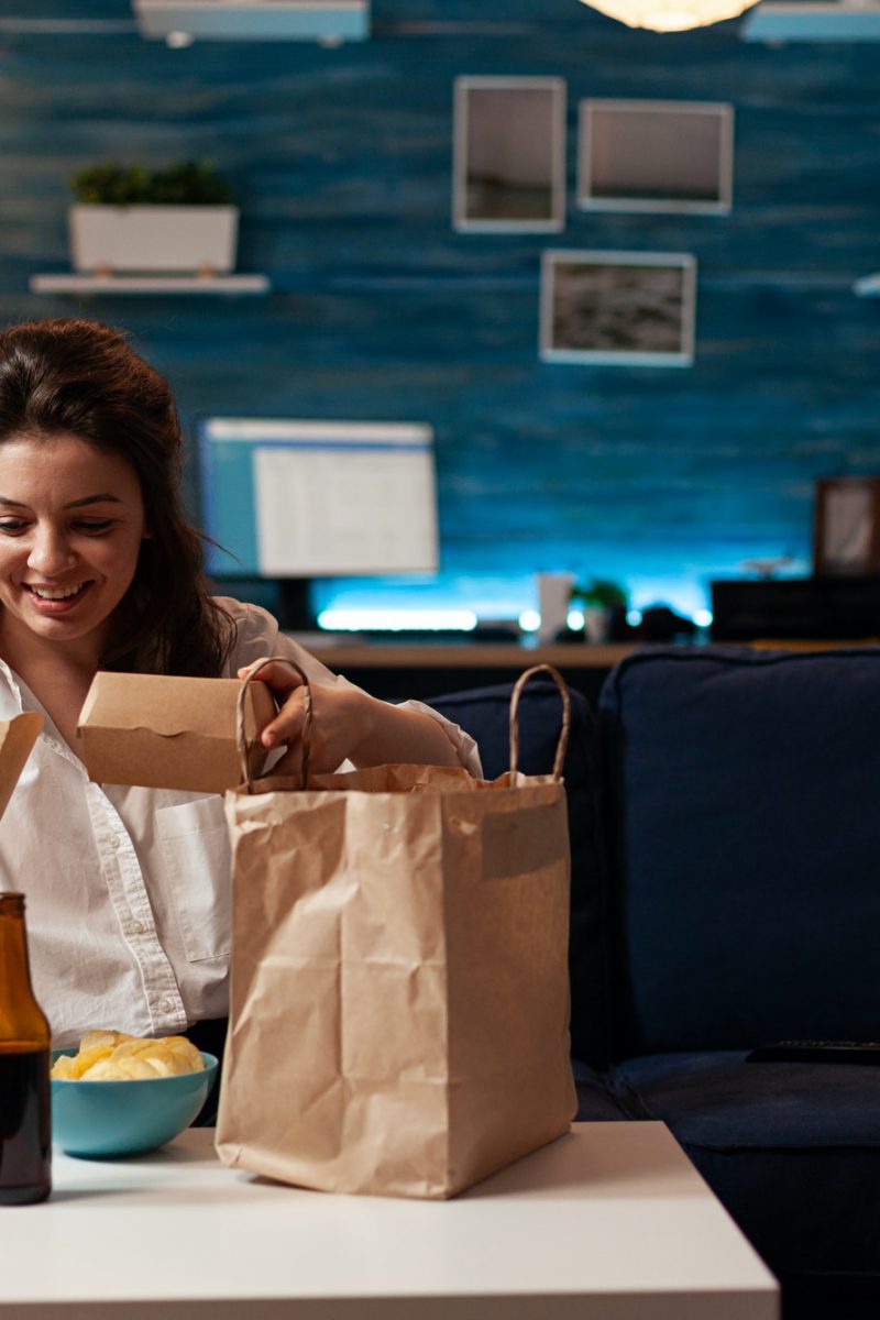 Smiling cheerful woman unpacking tasting fast-food home delivered sitting on couch